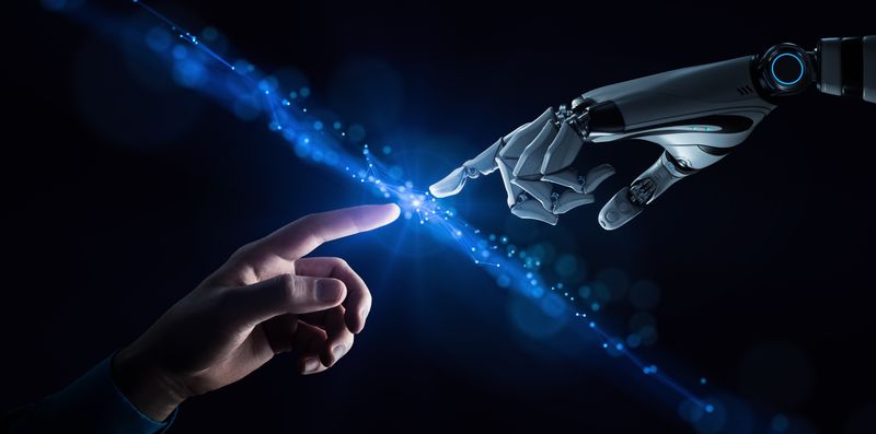 A digital conceptual photo showing a human hand extending into the middle of the image from the bottom left, while a robotic hand extends to the middle from the upper right hand corner. Both hands have their index fingers extended and are about to touch. There is a blue line of energy in the negative space between their fingers cutting diagonally across the image from the top left to the bottom right. 
