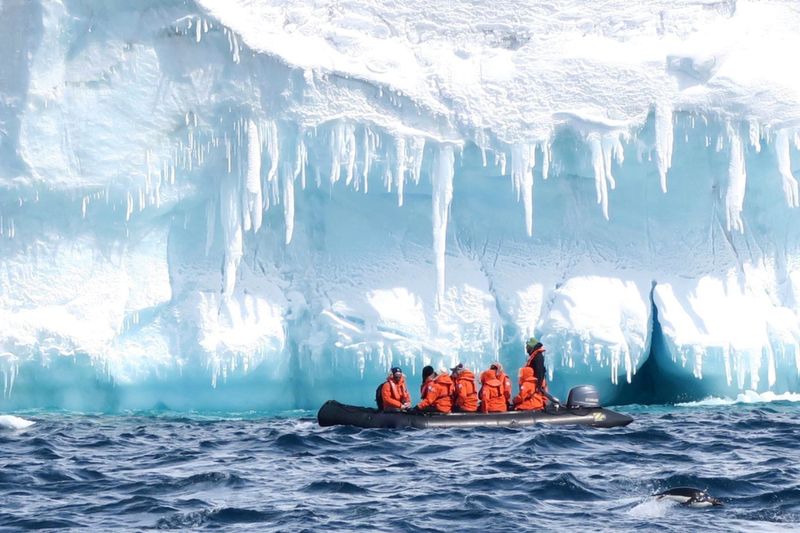 A boat of scientists in orange sailing next to a ice glacier in Antarctica