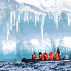 A boat of scientists in orange sailing next to a ice glacier in Antarctica