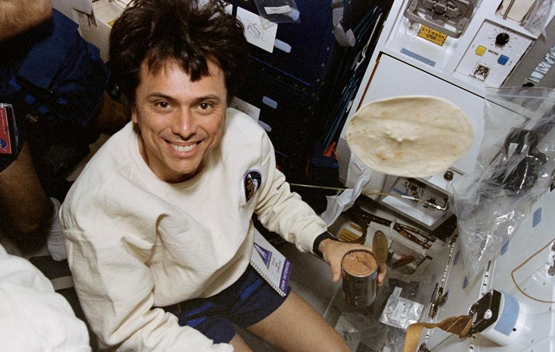 astronaut is smiling at the camera holding a bean can with a tortilla floating around
