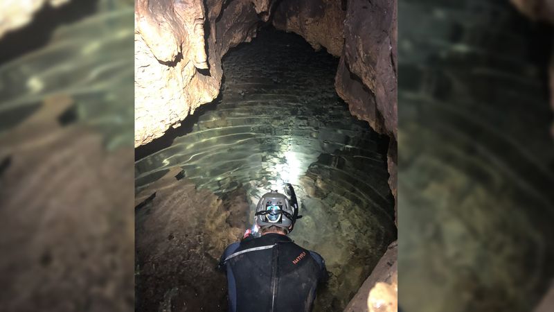A photo showing the back of a man wearing a wetsuit, a hardhat with a torch and a snorkel crouching by an underground stream in a cave. The water is clear and shallow, showing a rocky floor below the surface.  
