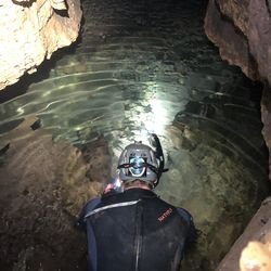 A photo showing the back of a man wearing a wetsuit, a hardhat with a torch and a snorkel crouching by an underground stream in a cave. The water is clear and shallow, showing a rocky floor below the surface.  