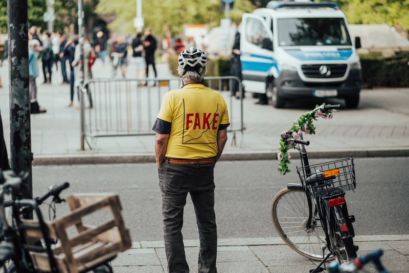 A photo of a man wearing a yellow t-shirt standing on a road side looking at other people walking in the background. The man has his back to the camera and is wearing a bike helmet. His t-shirt has the word "FAKE" written over a graph showing a rising trend. 