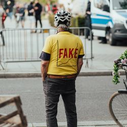 A photo of a man wearing a yellow t-shirt standing on a road side looking at other people walking in the background. The man has his back to the camera and is wearing a bike helmet. His t-shirt has the word "FAKE" written over a graph showing a rising trend. 