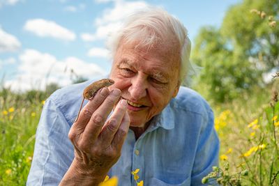 David Attenbourough smiling as he holds up a tiny harvest mouse to his face