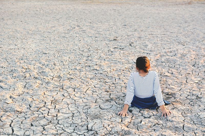 young girl sitting on cracked, dry ground facing away from the camera