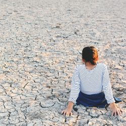 young girl sitting on cracked, dry ground facing away from the camera