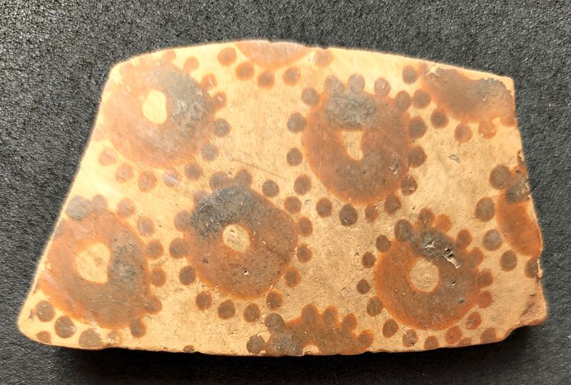 A pottery shard with around six crudely painted cirlces with dots around them to look like flowers 
