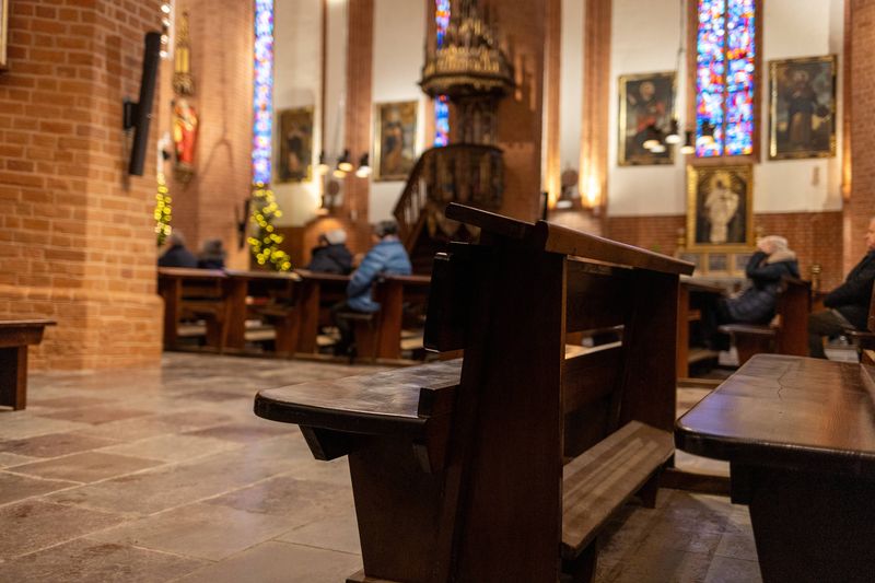 A photo showing two empty church pews in a catholic church. In the background, a column of pews with some people sat among them is visible, though blurred. The church is richly decorated with stain glass windows in the background. 
