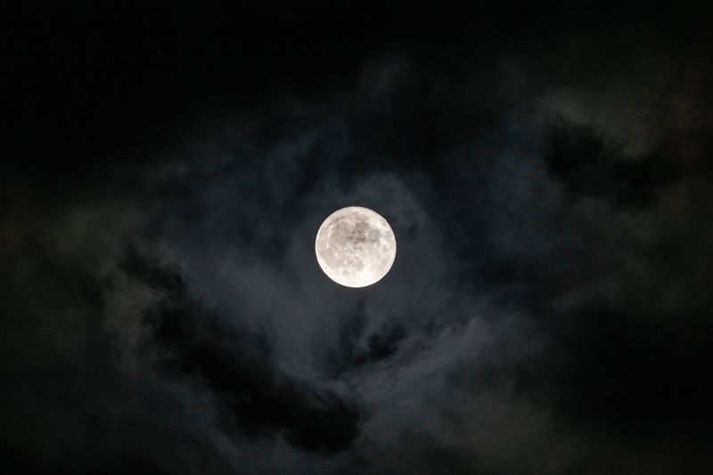 A photo taken of the full moon in a cloudy night sky. The moon itself is framed in the picture's center with darker and lighter clouds surrounding it.