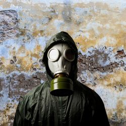 A photo of someone wearing a gas mask and a green plastic-like suit covering their body. They are standing looking directly at the camera with their back to a weathered, stained wall. 