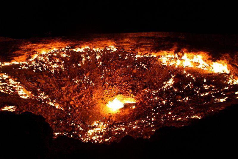 A photo taken at night of the crater. The bowl-like shape of the pit has a bright flame visible at its center, while the surrounding earth has smoldering patches. There are larger flames at the crater's edge which look like they're highlighting its contours.  of 