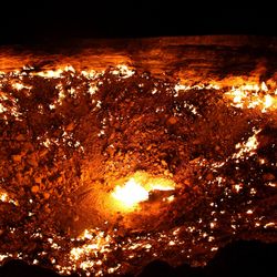 A photo taken at night of the crater. The bowl-like shape of the pit has a bright flame visible at its center, while the surrounding earth has smoldering patches. There are larger flames at the crater's edge which look like they're highlighting its contours.  of 