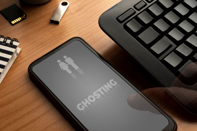 A photo showing someone's smart phone on a table next to a black keyboard. The phone has a dark screen on it with "GHOSTING" appearing in capital letters in its middle. Above this is the outline of a female and male figure standing next to one another. There is also a small memory drive and the corner of a notebook visible to the left of the phone.