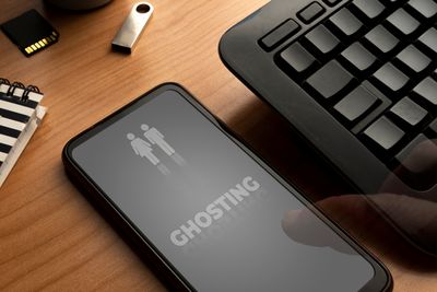 A photo showing someone's smart phone on a table next to a black keyboard. The phone has a dark screen on it with "GHOSTING" appearing in capital letters in its middle. Above this is the outline of a female and male figure standing next to one another. There is also a small memory drive and the corner of a notebook visible to the left of the phone.