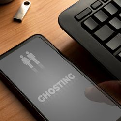 A photo showing someone's smart phone on a table next to a black keyboard. The phone has a dark screen on it with "GHOSTING" appearing in capital letters in its middle. Above this is the outline of a female and male figure standing next to one another. There is also a small memory drive and the corner of a notebook visible to the left of the phone.