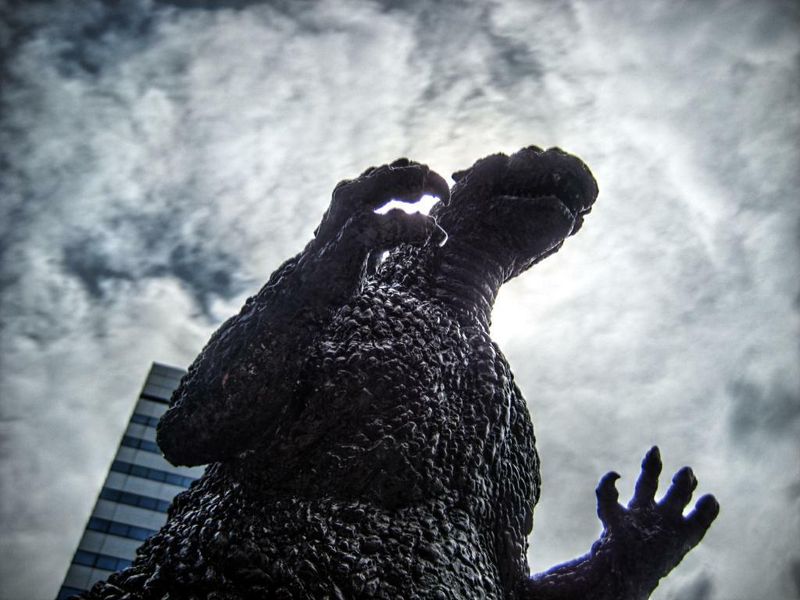godzilla statue photographed from below with a cloudy sky and a high-rise building in the background