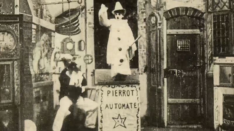 A still frame image showing a scene from Gugusse and the Automaton. In the center of the shot is a boy dressed as a clown standing on a pedestal with the word "Pierrot Automate" written on it. There is a star under the words. The boy is wearing a pointed clown hat, a long white coat with large bubble-like buttons and his face appears to have been painted white too. He is holding a stick. To the right is a man with a large moustache cranking a leaver that's attached to the clown's pedestal. The background depicts a painted screen of an old workshop, with bellows, a grandfather clock and an old door.  