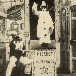 A still frame image showing a scene from Gugusse and the Automaton. In the center of the shot is a boy dressed as a clown standing on a pedestal with the word "Pierrot Automate" written on it. There is a star under the words. The boy is wearing a pointed clown hat, a long white coat with large bubble-like buttons and his face appears to have been painted white too. He is holding a stick. To the right is a man with a large moustache cranking a leaver that's attached to the clown's pedestal. The background depicts a painted screen of an old workshop, with bellows, a grandfather clock and an old door.  