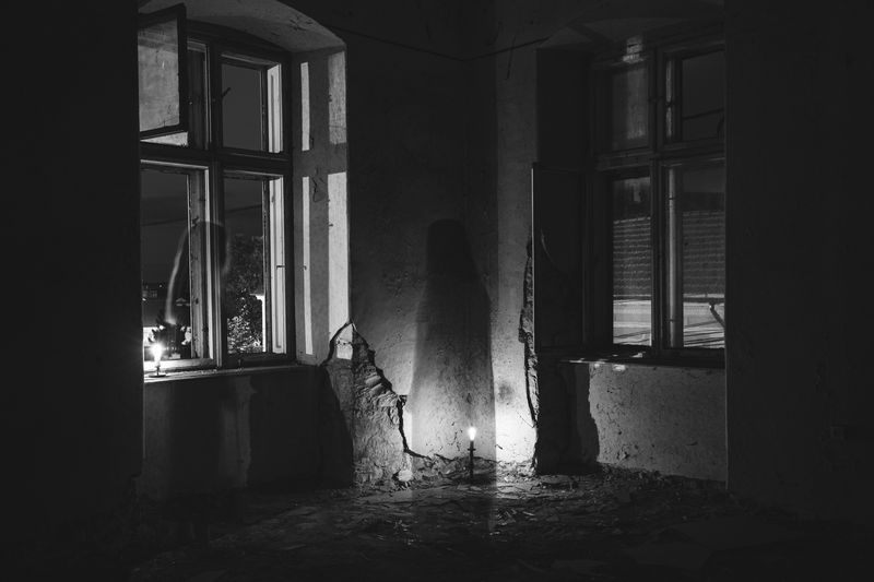 A black and white image of the corner of an abanonded room with a singl;e lightbulb on the floor and, if you squint, the shadow of a ghostly figure by the window