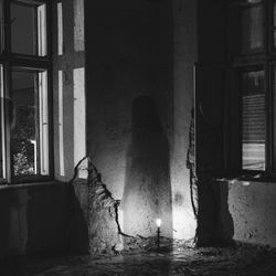 A black and white image of the corner of an abanonded room with a singl;e lightbulb on the floor and, if you squint, the shadow of a ghostly figure by the window