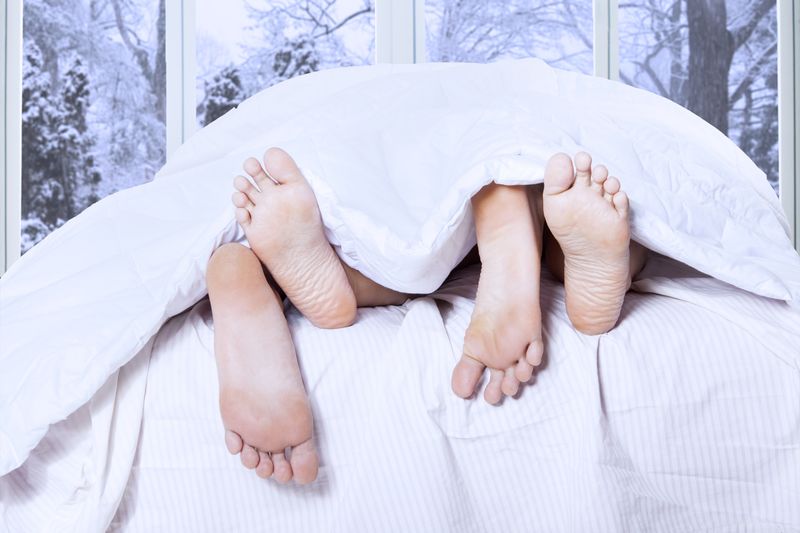 shot from the end of a bed with two people's feet sticking out under the duvet; there's a window behind the bed showing a wintry snowy scene