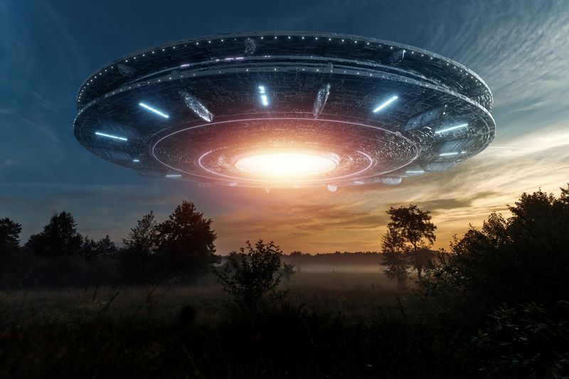 An image showing a digitally created UFO looming over some trees on what looks like a misty morning. The UFO looks like two large discs put together - like and Oreo - and it has a large bright light in the center of its underside. 