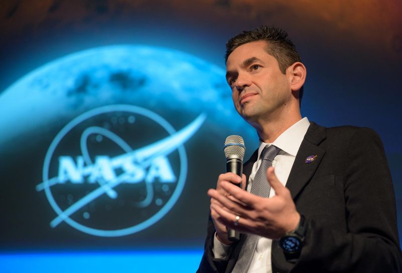 Isaacman, a white man in his 50s in from of a screen with the moon and a nasa logo holding a mike
