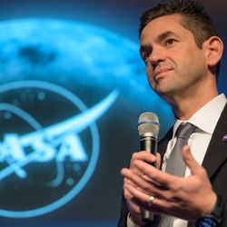 Isaacman, a white man in his 50s in from of a screen with the moon and a nasa logo holding a mike