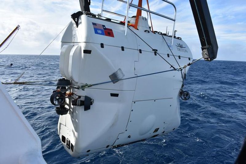 A photo showing a white submersible being held ahead of being put into the water. The vessel is shaped like a suit-case or brief-case and is white, showing distinct panels along its sides. There is a railing visible at the top. 