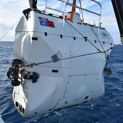 A photo showing a white submersible being held ahead of being put into the water. The vessel is shaped like a suit-case or brief-case and is white, showing distinct panels along its sides. There is a railing visible at the top. 