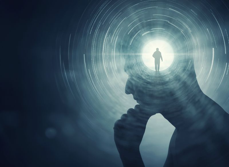 A conceptual image showing man's silhouette against a darker backdrop. The man is position on profile, facing to the left and appears to be rubbing his chin in thought. The center of his head shows a bright circle with a smaller silhouette of a man walking in it. This small circle is radiating light that gives the man's main profile definition against the dark surrounding him.