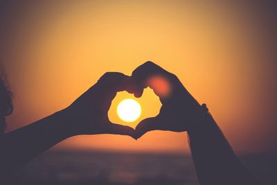 silhouette of two people's hands forming a heart symbol around the setting sun