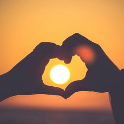 silhouette of two people's hands forming a heart symbol around the setting sun