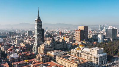 A skyline photo showing Mexico city sprawling out. In the center of the image is a tall tower building surrounded by smaller, flat roofed buildings. 