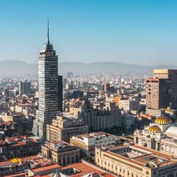A skyline photo showing Mexico city sprawling out. In the center of the image is a tall tower building surrounded by smaller, flat roofed buildings. 