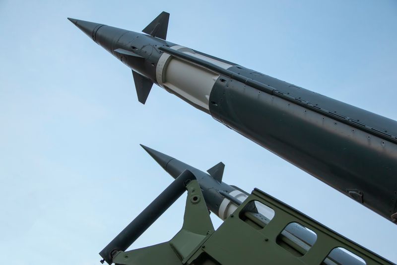 A photo taken under two defense missiles pointing at the sky. The missiles enter the frame from the bottom right and extend to the top left. They are a dark green color with a white band towards their top. 