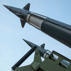 A photo taken under two defense missiles pointing at the sky. The missiles enter the frame from the bottom right and extend to the top left. They are a dark green color with a white band towards their top. 