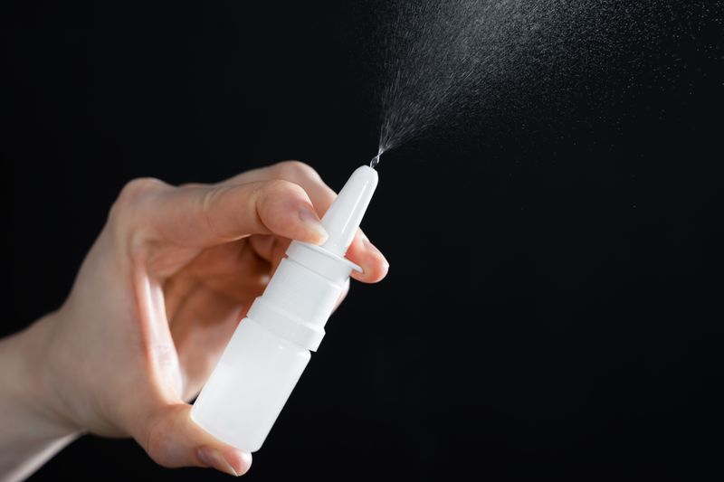 A photo showing a person's hand holding an unlabeled nasal spray. The spray is uniform white and it is ejecting a mist into the air. It is framed against a black background. 