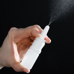 A photo showing a person's hand holding an unlabeled nasal spray. The spray is uniform white and it is ejecting a mist into the air. It is framed against a black background. 