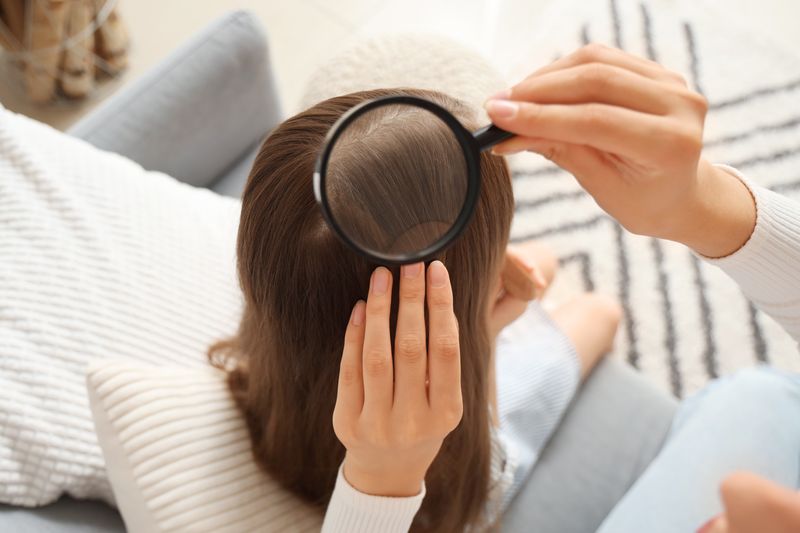 A photo of looking down on the woman examining the back of a girl's head with a magnifying glass. The girl is in the center of the shot and the woman is sitting to the right, just out of sight. She has her left hand on the girl's hair and is holding a magnifying glass in right to examine it. They are sitting on a sofa with white pillows.