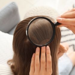A photo of looking down on the woman examining the back of a girl's head with a magnifying glass. The girl is in the center of the shot and the woman is sitting to the right, just out of sight. She has her left hand on the girl's hair and is holding a magnifying glass in right to examine it. They are sitting on a sofa with white pillows.