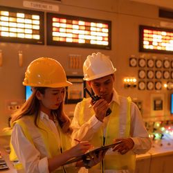 Two young nuclear engineers stand in a control room surrounded by panels with buttons and lights. They are wearing hard hats and high visibility jackets. The female engineer is standing on left and examining a clipboard while the male engineer to her right is talking into a walkie-talkie.  