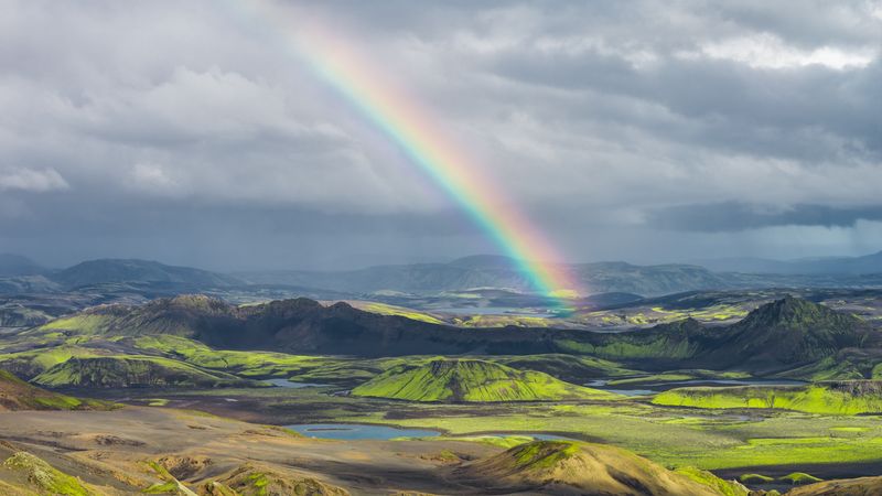 Rugged landscape with green hills, volcanic soil, scattered water bodies, and a vivid rainbow under a cloudy sky.