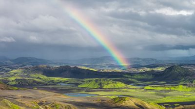Rugged landscape with green hills, volcanic soil, scattered water bodies, and a vivid rainbow under a cloudy sky.
