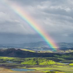 Rugged landscape with green hills, volcanic soil, scattered water bodies, and a vivid rainbow under a cloudy sky.