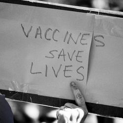 RFK jr against a black and white backdrop of a demonstration sign reading "vaccines save lives"