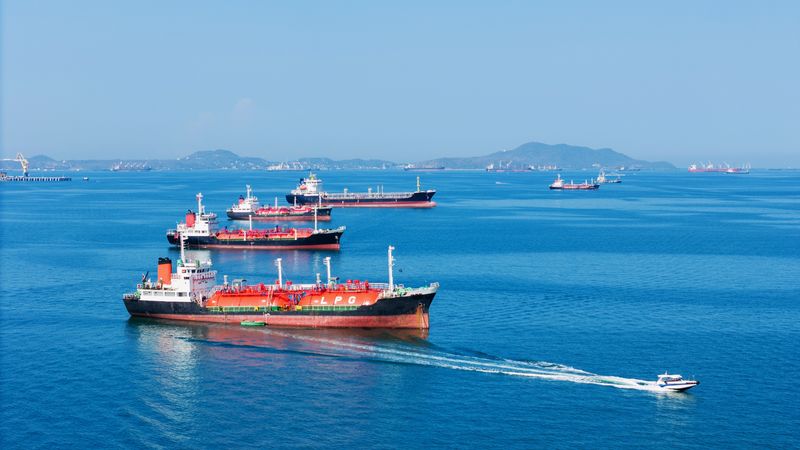 A photo showing several oil and gas vessels anchored in the Strait of Hormuz. They are lined up, fading into the distance on pristine blue water. There is a smaller patrol boat passing the forward most vessel. 