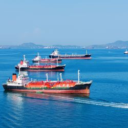 A photo showing several oil and gas vessels anchored in the Strait of Hormuz. They are lined up, fading into the distance on pristine blue water. There is a smaller patrol boat passing the forward most vessel. 