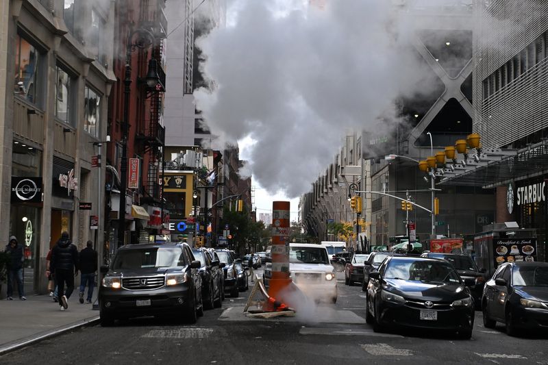 A photo showing a busy street with cars lining up either side of a pipe with plumes of steam floating above it. 